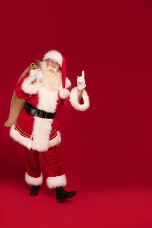 Full length photo of real Santa Claus carrying gifts, posing on red studio background. Christmas is coming! A lot of copy space. Xmas time. Holiday discounts concept.の写真素材