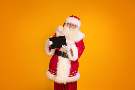 Cheerful Santa Claus showing digital tablet to the camera, posing on yellow studio background. A lot of copy space. Xmas discount, sale concept. Christmas time.の写真素材