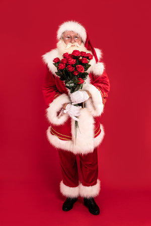 Photo of real Santa Claus standing on red studio background with roses in hands, looking at the camera. Xmas date. Christmas sale, discount concept.の写真素材