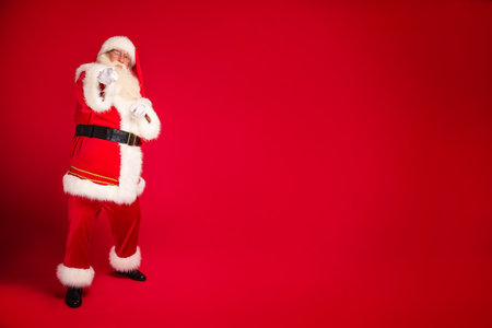 Full length photo of real Santa Claus annoucing big xmas sale on a red studio background, pointing to the camera. Merry Christmas and Happy New Year! A lot of copy space for your text.の写真素材