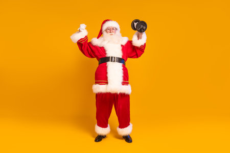 Full length photo of real Santa Claus doing fit gym exercise. Christmas time. Xmas vibes. A lot of copy space on the yellow studio background.の写真素材
