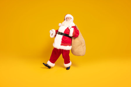 Merry Christmas and a happy new year! Real Santa Claus carrying heavy sack with gifts, standing on yellow studio background. Xmas time. A lot of copy space.の写真素材