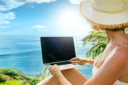 Digital nomad working on the tropical beach with laptop computer. Woman in bikini and summer hat using technology for online shopping on vacation.の写真素材