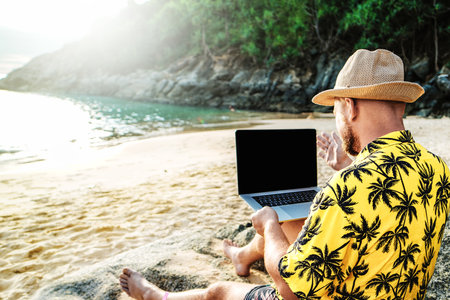 Man in the summer hat , a businessman, digital nomad working with laptop on the rocky beach.  Freelancer. Tropics. Summer vibes.の写真素材
