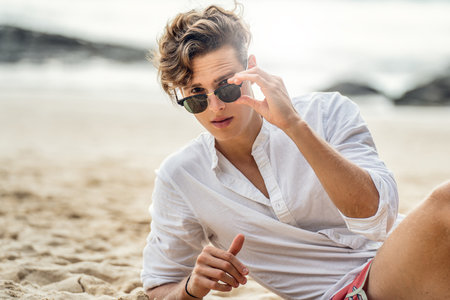 Portrait of handsome young italian man relaxing at the beach, wearing white shirt and fashionable sunglasses. Summer time.の写真素材
