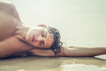 Beauty portrait of handsome young male model relaxing at the beach, lying on the sand. Summer vibes.の写真素材