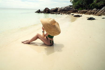 Photo fo a woman enjoying sunbath at tropical beach, sitting on the sand, wearing big summer hat. Tanned girl relaxing on vacation. tourists. island.の写真素材