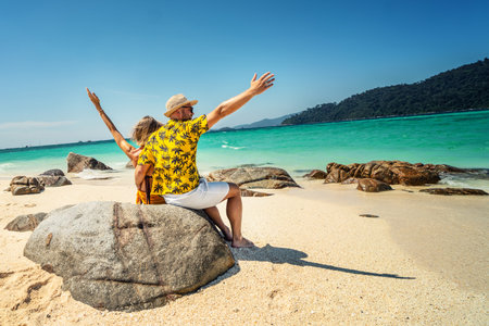 Happy Couple Enjoying Beautiful seascape, sitting with hands up on the rock, on the tropical beach. Travel Vacation, real people Lifestyle Concept. back view.の写真素材