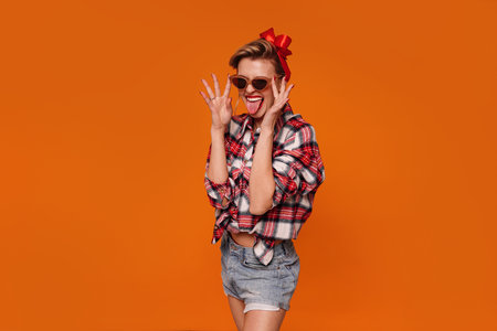Photo of crazy pinup girl in checkered shirt and short jeans smiling and showing tongue out. Retro vintage 50s style, studio shot isolated on orange background. A lot of copy space.の写真素材