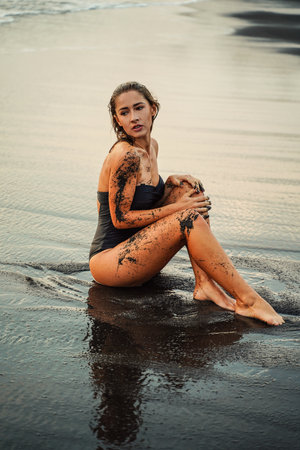 Sexy woman in swimsuit sitting on the black beach. Tanned woman with perfect body shape. Relax and natural body treatment.の写真素材