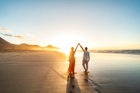 Lovely, romantic couple walking on sunset beach, enjoying evening light, relaxing on tropical summer vacation. Honeymoon. love. back view. Woman wearing orange maxi dress, man in linen pant and white shirt.の写真素材