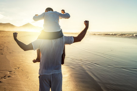 Happy father and son enjoying time at tropical beach during family vacation, playing and having fun together. back view. superheroes. boys power. tourism. Travelers.の写真素材