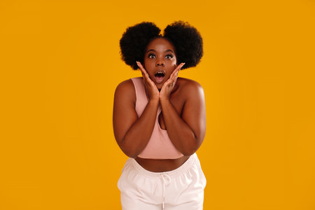 Shocked concerned african-american woman with afro hairstyle looking at the camera and holding palms on face from surprise posing against orange studio background. A lot of copy space.の写真素材