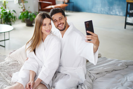 Cheerful and happy couple taking selfie in bedroom, having fun together. Man and woman sitting on the bed, looking at the phone screen.の写真素材