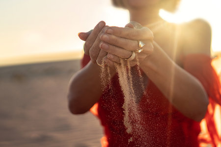 Elegant woman with sand falling through her hands on the beach at sunset time. golden light. concept photo.の写真素材