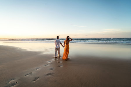 Romantic couple walking on sunset beach, enjoying evening light, relaxing on tropical summer vacation. Honeymoon. love. back view. Woman wearing orange maxi dress, man in linen pant and white shirt.の写真素材