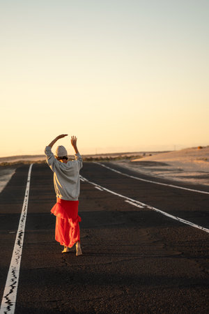 adventure concept. Back view of a hippie woman walking alone and barefoot on the highway, dancing. sunset light. summer vibe.の写真素材