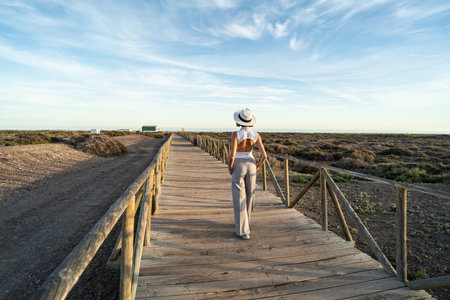 Stylish woman in white hat walking on the wooden promenade on the sea shore, relaxing , enjoying sunny day. Summer vacation. Space for text. Back view. Full length photo.の写真素材