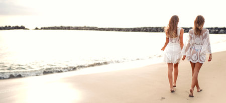 Back view of two blonde women walking together on the sandy beach, holding hands. friendship concept. Full length photo.の写真素材