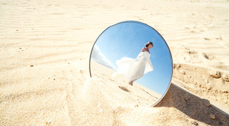 Conceptual photo, reflection in the mirror of attractive blond woman in long white wedding dress posing in desert. Sandy dunes and blue sky.の写真素材