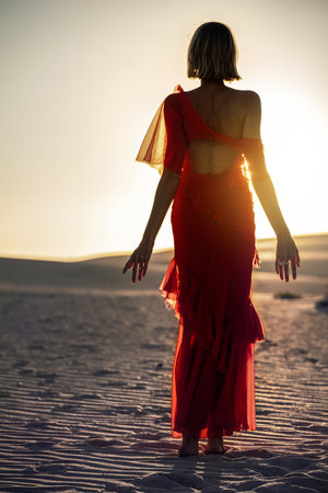 Back view of a romantic woman in a red maxi dress posing in the sand desert at sunset light. Full length photo.の写真素材