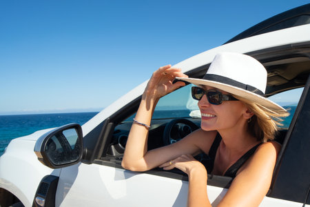 Happy woman holding summer hat outside open window in car. People lifestyle. Traveler on road trip. Blue sky.の写真素材