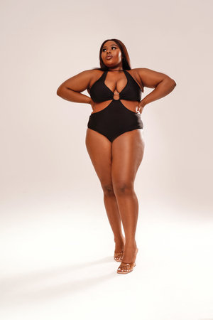 Full length photo of beautiful, dark skinned woman posing on light gray studio background, wearing black fashionable swimwear. Body positive, conscious concept. Afro hairstyle. copy space. selflove.の写真素材