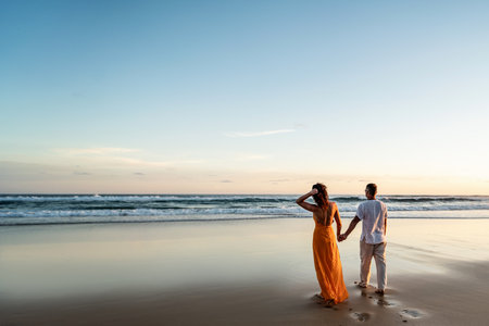 Romantic couple walking on sunset beach, enjoying evening light, relaxing on tropical summer vacation. Honeymoon. love. back view. Woman wearing orange maxi dress, man in linen pant and white shirt.の写真素材