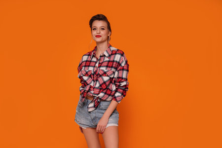 Fashion photo of happy, beautiful woman in short jeans and checkered shirt. Girl posing on orange studio background. A lot of copy space.の写真素材