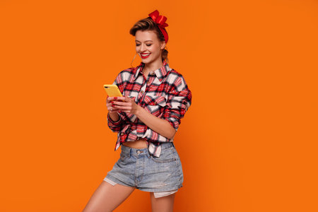 Studio Shot Of Smiling, Happy, Young Woman Using Mobile Phone, scrolling social media. Orange studio background. A lot of copy space. Internet addiction.の写真素材