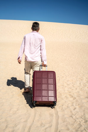 Looking for the sea and relax. Traveling and summer vacation concept. Back view of a man walking at the sandy beach with suitcase. Full length photo. blue sky. tourism.の写真素材