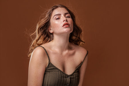 Super natural, beautiful girl looking at the camera. Attractive woman with freckles on her face and body and long wavy hair. Brown studio background with a lot of copy space.の写真素材