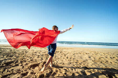 Superhero child having fun on the sandy, sunset beach, wearing red mask and cape, running. Summer vacation concept. Copy space. Kids power.の写真素材