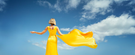 Fashionable blonde woman in yellow dress posing on the blue sky background, back view. A lot of copy space. Colors.の写真素材