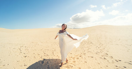 Calm woman in fashionable, maxi white wedding dress posing on the desert, dancing in sunny light, A lot of copy space. Bride to be.の写真素材