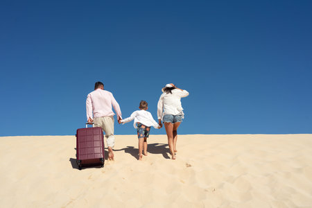 Mother and father with their son walking on the sandy beach, dunes with suitcases. Concept of the family vacation and tourism. blue sky. copy space. back view.の写真素材