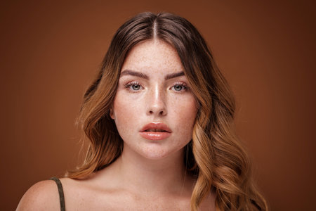 Super natural, beautiful girl looking at the camera. Attractive woman with freckles on her face and long wavy hair. Brown studio background.の写真素材