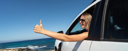 Blonde traveler woman smiling happy with thumb up doing ok sign sitting at the car, enjoying sunny day at summer vacation. blue sky. Real people emotions. tourists. car hire.の写真素材