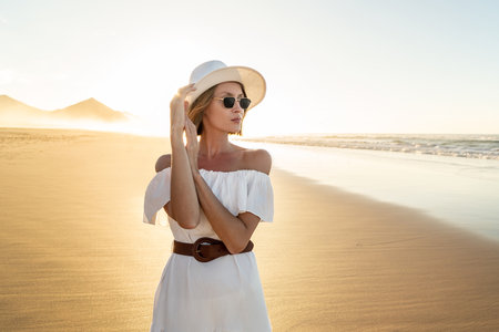 Fashionable woman in white dress and summer hat posing on the sandy beautiful beach at sunset light.の写真素材