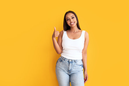 Young woman wearing casual clothes, white top and jeans, showing okay sign with fingers, smiling to the camera. Yellow studio background with copy space. Real people emotions. Happiness and fun.の写真素材