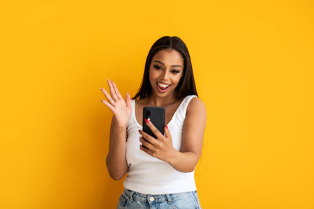Young happy woman wearing basic clothes smiling to the cellphones empty screen, posing over yellow studio background. Real people emotions. copy space.の写真素材