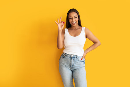 Young woman wearing casual clothes, white top and jeans, showing okay sign with fingers, smiling to the camera. Yellow studio background with copy space. Real people emotions. Happiness and fun.の写真素材
