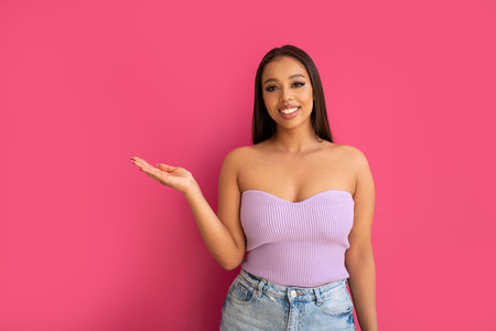 Cheerful young lady in fashionable top pointing at copy space on pink studio background. Smiling woman showing some advertisement or announcement, looking at the camera.の写真素材