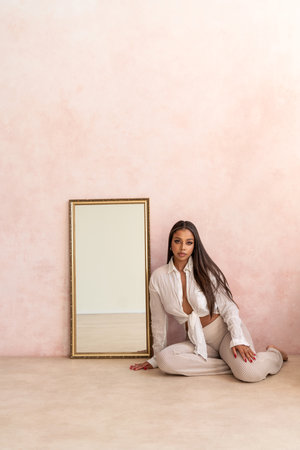 Attractive woman with long dark hair wearing white fashionable shirt, sitting on the floor next to the vintage mirror, looking at the camera. A lot of copy space. Light pastel tone. Full length photo.の写真素材