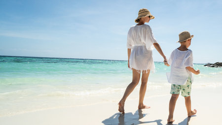 Back view of happy mother and son walking, relaxing on the tropical sandy beach, Enjoying life. Summer day. Vacation. Travel.の写真素材