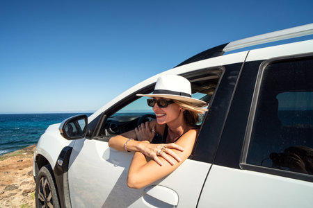 Happy woman in white hat on a road trip traveling by rental car . Adventure lifestyle, vacations vibes. Smiling female looking out of the car window.の写真素材