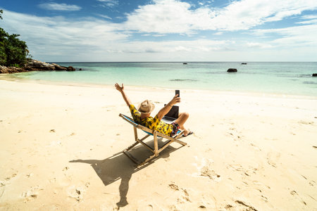 Man in the summer hat , a businessman, digital nomad working with laptop and mobile phone on the tropical beach. Freelancer. Tropics. Summer vibes.の写真素材