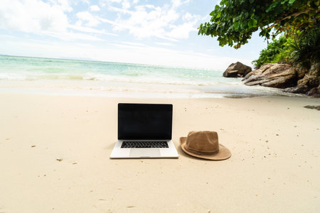 Conceptual photo of modern laptop and summer hat on the sandy beach - travel background. Digital nomad lifestyle.の写真素材