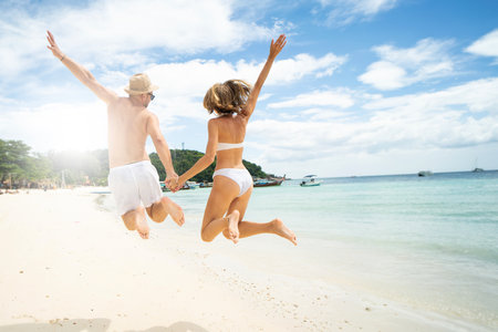 Full body back view of happy couple in swimwear jumping on sandy tropical beach, holding hands, having fun together. Summer vibes. Travel concept.の写真素材