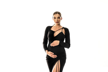 Pregnancy concept. Woman posing in white studio background wearing black dress, holding her pregnant belly, looking at the camera. A lot of copy space.の写真素材